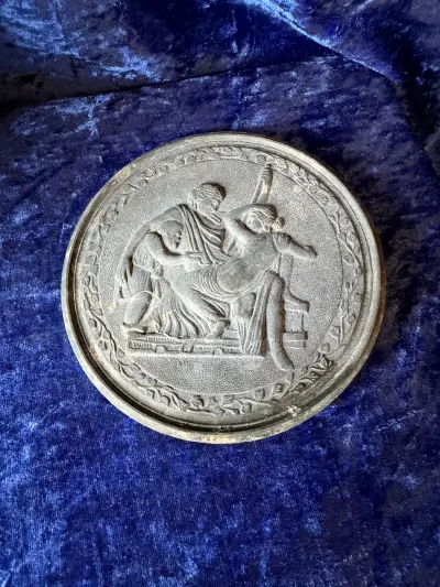 Rundt metalrelief (vægplatte) med et klassisk motiv, der ligner Bertel Thorvaldsens værker eller klassiske græske/romerske mytologiske motiver (såsom Alderdommen eller Geras og Hebe), udført i mørkt m
